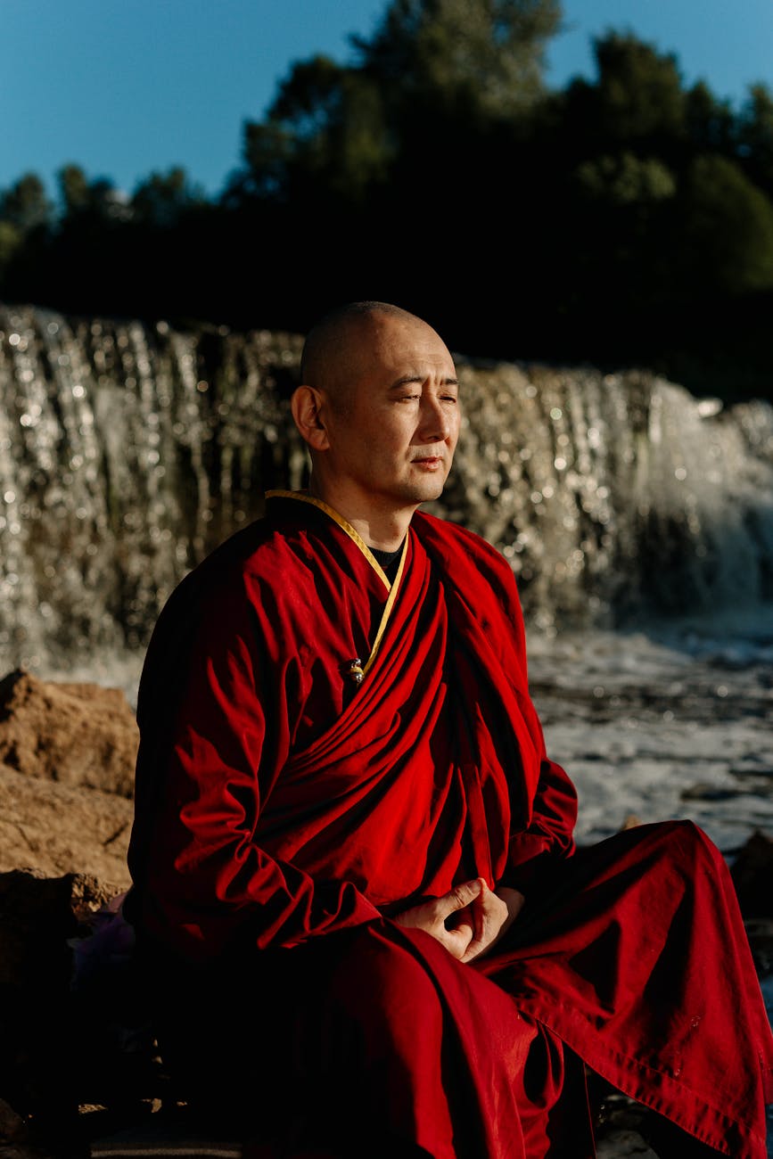 man in red robe sitting on rock near water falls